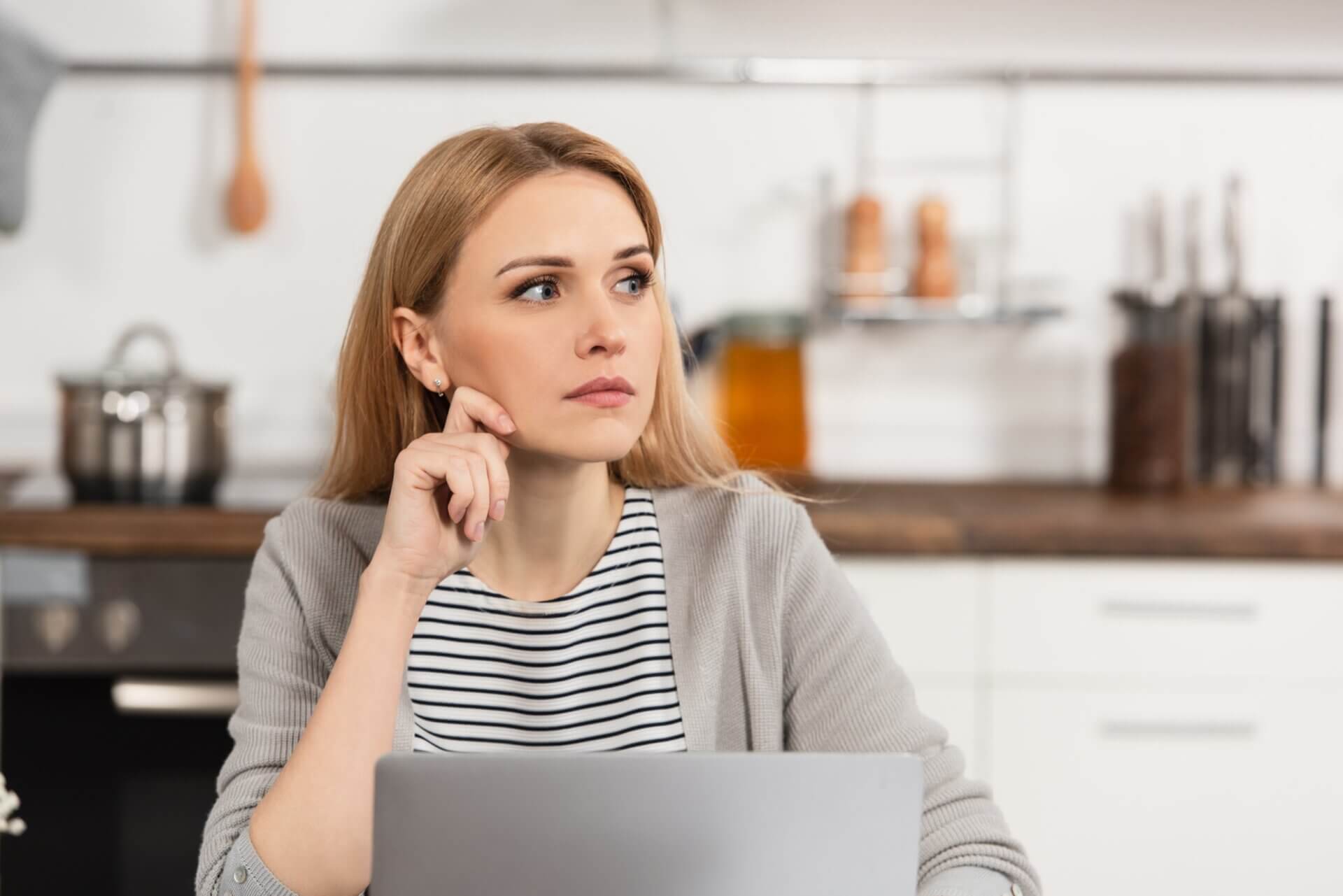 Mujer pensativa mientras trabaja en el ordenador desde la cocina