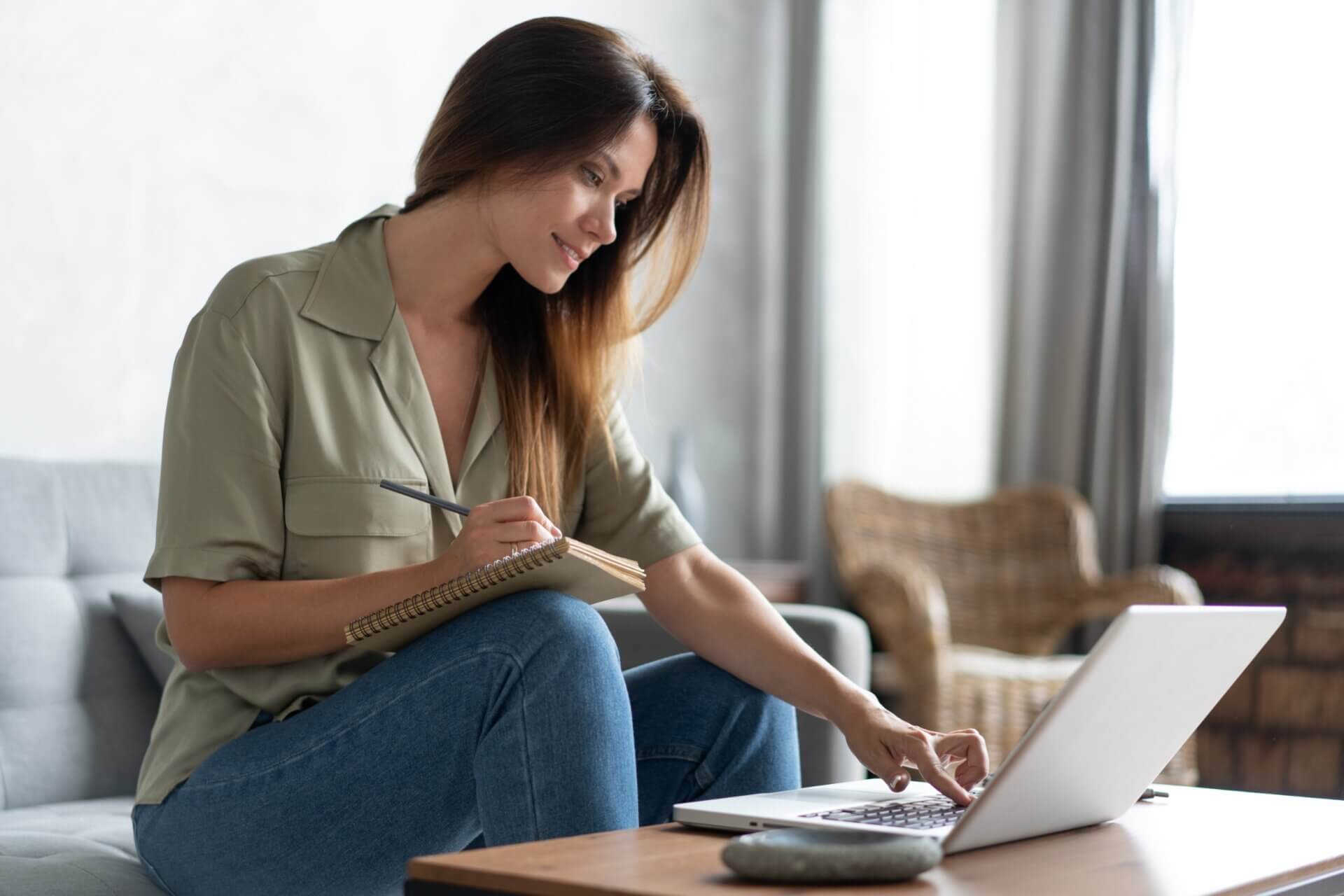 Mujer toma notas en una libreta mientras trabaja en el ordenador
