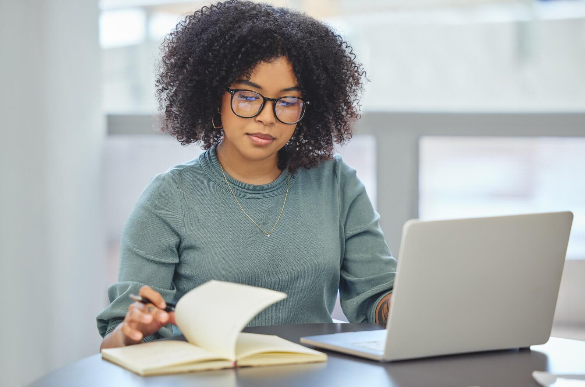 Mujer con gafas escribe libreta mientras trabaja en el ordenador
