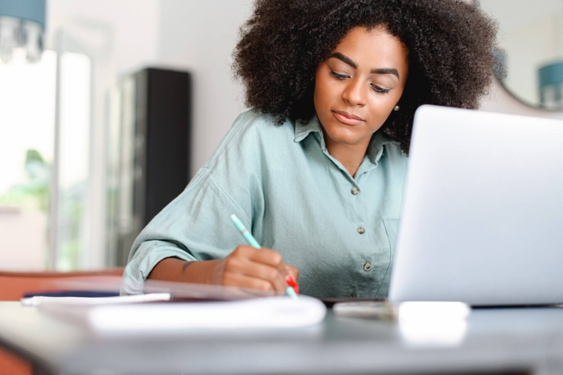 Mujer con cabello rizado toma notas mientras trabaja en el ordenador